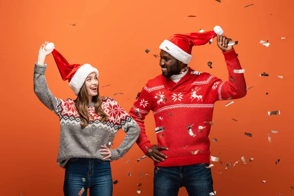 Heureux couple interracial dans santa chapeaux et chandails de Noël en regardant les uns les autres sous confettis sur fond orange — Photo de stock