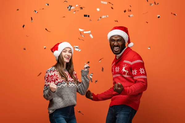 Glückliches gemischtrassiges Paar in Weihnachtsmützen und Weihnachtspullis unter Konfetti auf orangefarbenem Hintergrund — Stockfoto