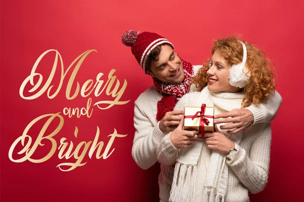 Heureux couple en tenue d'hiver tenant cadeau de Noël sur rouge avec un lettrage joyeux et lumineux — Photo de stock