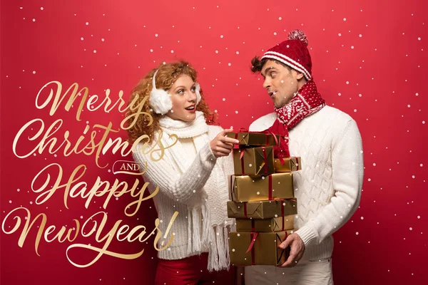 Heureux couple en tenue d'hiver tenant cadeaux de Noël sur rouge avec joyeux Noël et bonne année lettrage — Photo de stock