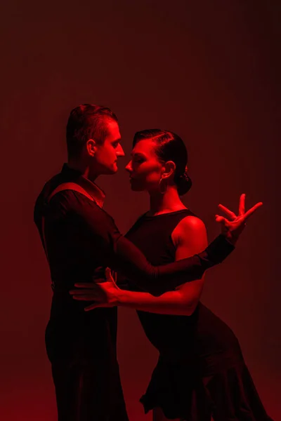 Apasionada pareja de bailarines en ropa negra realizando tango sobre fondo oscuro con iluminación roja - foto de stock