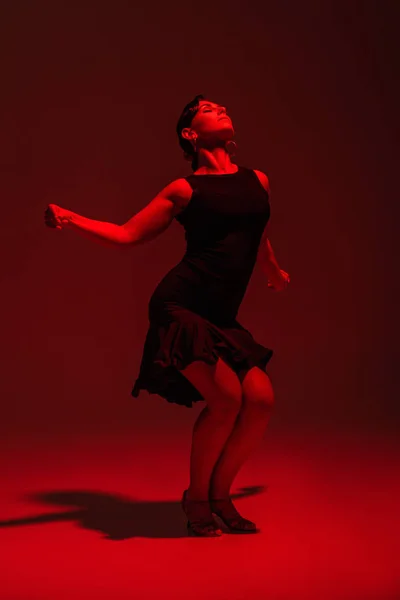 Sensual dancer in black dress performing tango on dark background with red lighting — Stock Photo