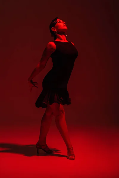 Beautiful dancer in black dress performing tango on dark background with red lighting — Stock Photo