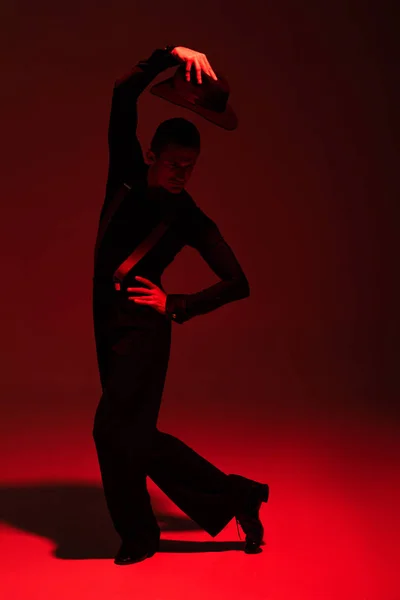 Elegante Tänzerin mit Hut über dem Kopf, während sie Tango auf dunklem Hintergrund mit roter Beleuchtung aufführt — Stockfoto