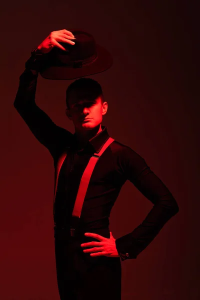 Expresiva bailarina de tango mirando a la cámara y sosteniendo el sombrero sobre la cabeza sobre fondo oscuro con luz roja - foto de stock
