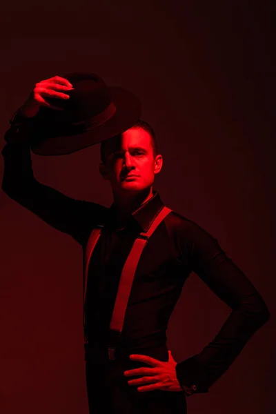 Hermosa bailarina de tango mirando a la cámara y sosteniendo el sombrero por encima de la cabeza sobre fondo oscuro con iluminación roja - foto de stock