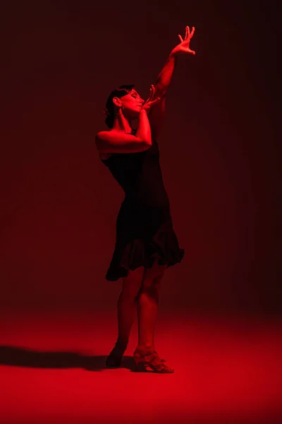 Élégante danseuse en robe noire exécutant le tango sur fond sombre avec éclairage rouge — Photo de stock
