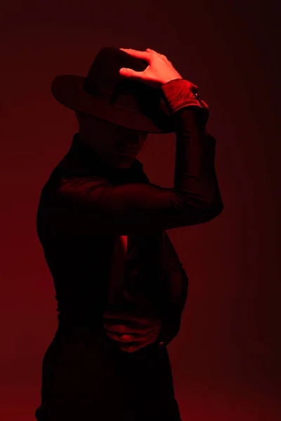Expressive dancer in black clothing and hat performing tango on dark background with red illumination — Stock Photo