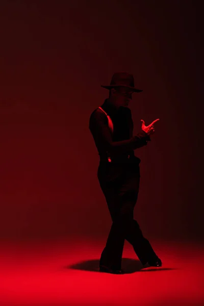 Elegante bailarina en ropa negra y sombrero realizando tango sobre fondo oscuro con iluminación roja - foto de stock