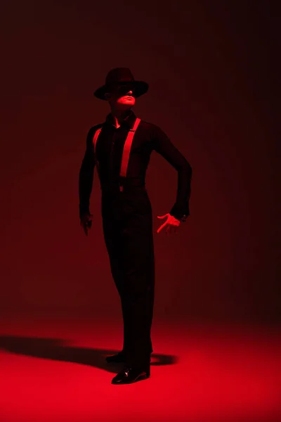 Sensual bailarina en ropa negra y sombrero realizando tango sobre fondo oscuro con iluminación roja - foto de stock