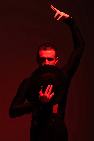 Bailarina apasionada mirando a la cámara y sosteniendo el sombrero cerca de la cara mientras realiza tango sobre fondo oscuro con iluminación roja — Stock Photo