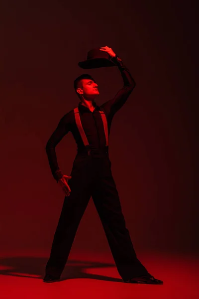 Стильный танцор, держащий шляпу над головой во время исполнения танго на темном фоне с красным освещением — стоковое фото