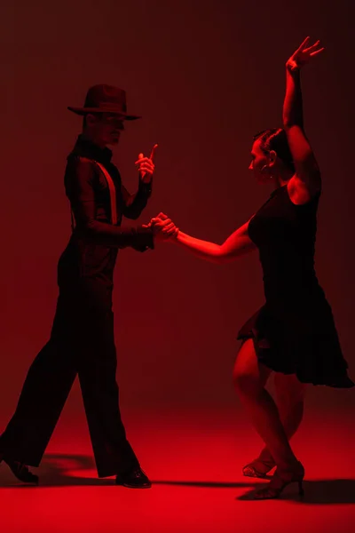 Элегантная пара танцоров в черной одежде, исполняющих танго на темном фоне с красной подсветкой — стоковое фото