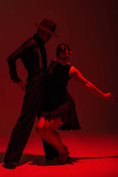 Apasionada pareja de bailarines en ropa negra realizando tango sobre fondo oscuro con iluminación roja - foto de stock