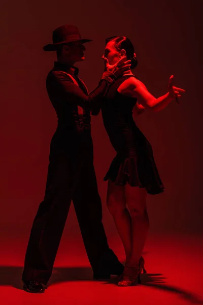 Expressive couple of dancers in black clothing performing tango on dark background with red lighting — Stock Photo