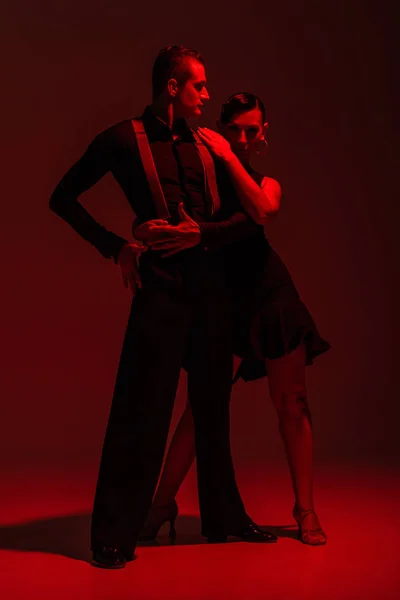 Apasionada pareja de bailarines en ropa negra realizando tango sobre fondo oscuro con luz roja - foto de stock