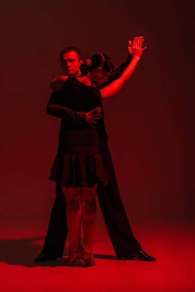 Élégant couple de danseurs en vêtements noirs exécutant tango sur fond sombre avec éclairage rouge — Photo de stock