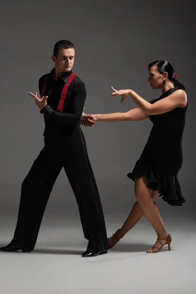 Apasionada pareja de bailarines en ropa negra realizando tango sobre fondo gris - foto de stock