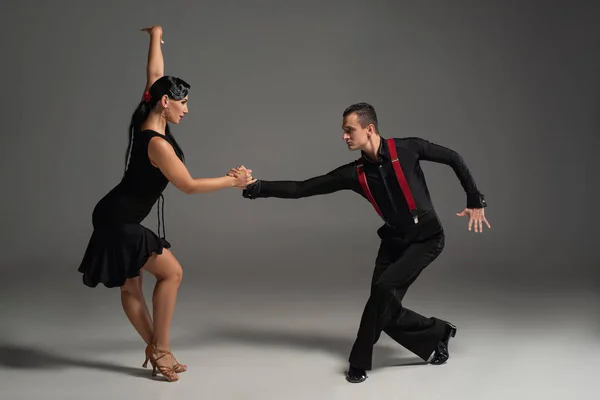 Стильні танцюристи в чорному одязі дивляться один на одного, танцюючи танго на сірому фоні — стокове фото