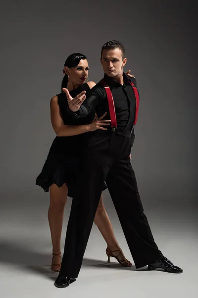 Apasionada pareja de bailarines en ropa negra realizando tango sobre fondo gris - foto de stock