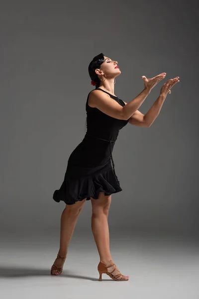 Elegante bailarina sensual en vestido negro realizando tango con los ojos cerrados sobre fondo gris - foto de stock