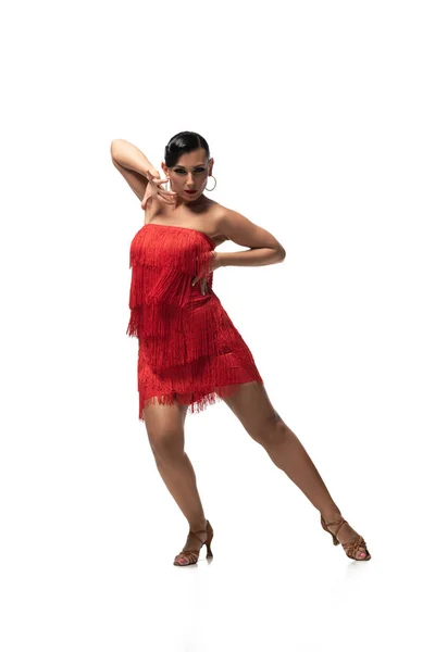 Привлекательный, страстный танцор в красном платье с бахромой, смотрящий в камеру во время исполнения танго на белом фоне — стоковое фото