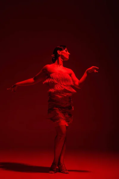 Красивая танцовщица в платье с бахромой, исполняющая танго на темном фоне с красной подсветкой — стоковое фото