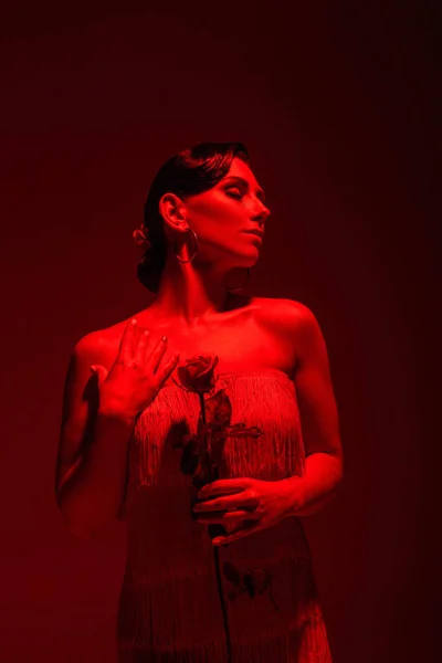 Елегантна танго танцюристка з закритими очима тримає червону троянду на темному фоні з червоним освітленням — стокове фото