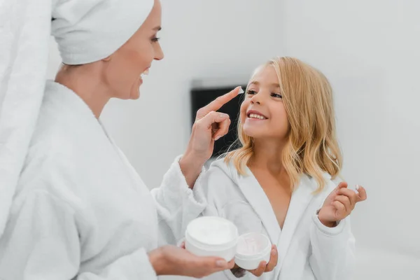 Fröhliche Mutter trägt kosmetische Creme auf sauberes Gesicht ihrer Tochter auf — Stockfoto