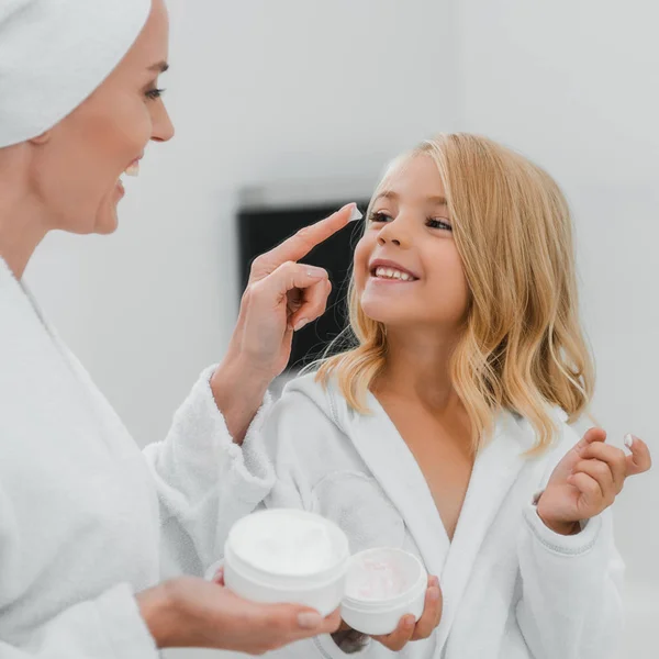 Mãe feliz aplicando creme cosmético no rosto limpo da filha — Fotografia de Stock