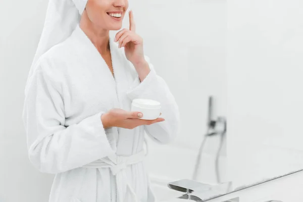 Vista ritagliata di donna sorridente applicare crema cosmetica sul viso pulito — Foto stock