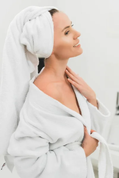 Cheerful woman in bathrobe touching neck in bathroom — Stock Photo