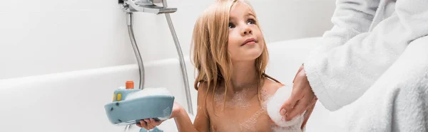Panoramic shot of cute kid holding rubber toy and looking at mother in bathroom — Stock Photo