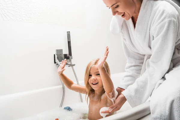 Joyeuse mère près de l'enfant avec les mains au-dessus de la tête prenant bain dans la baignoire avec mousse de bain — Photo de stock