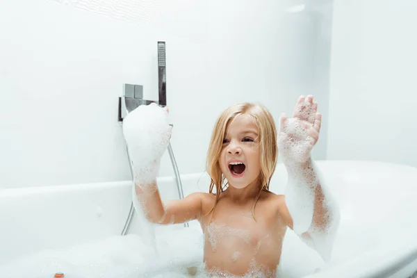 Lindo niño con la boca abierta y espuma de baño en las manos - foto de stock