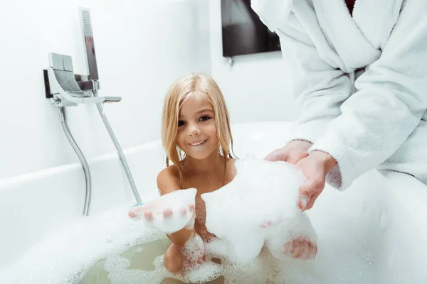 Criança feliz e mãe com espuma de banho a mãos — Fotografia de Stock