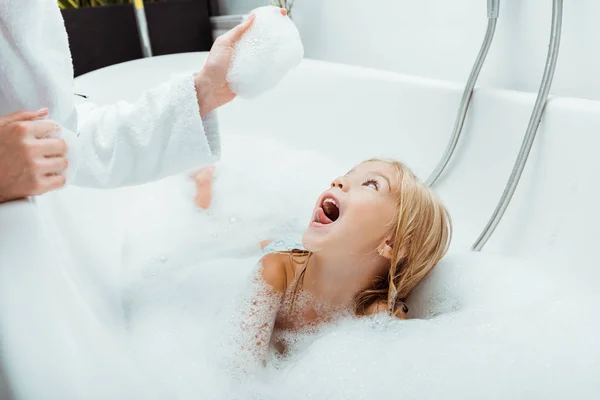 Aufgeregtes Kind blickt Mutter in Badeschaum im Badezimmer an — Stockfoto