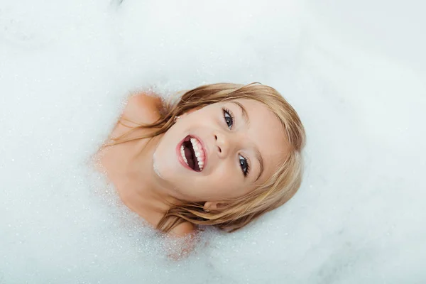 Vista superior de niño feliz tomando baño y mirando a la cámara en casa - foto de stock
