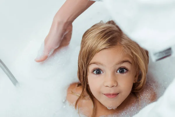 Vista aérea de un niño feliz tomando un baño y mirando la cámara cerca de la madre - foto de stock
