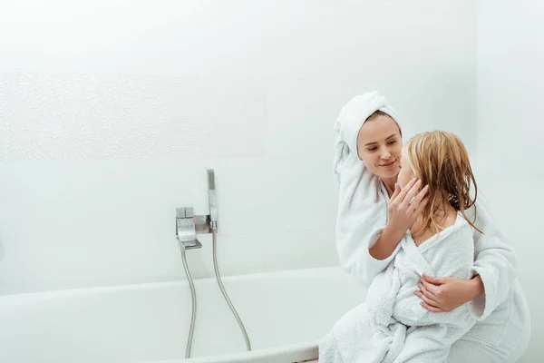 Feliz madre tocando la cara de la hija mientras está sentada en el baño - foto de stock