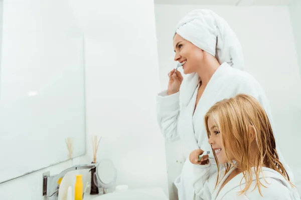 Cheerful mother and daughter in bathrobes brushing teeth near mirror — Stock Photo