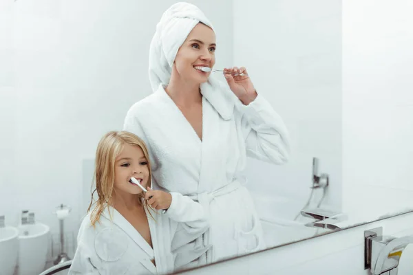 Feliz madre e hija en albornoces cepillarse los dientes cerca del espejo - foto de stock