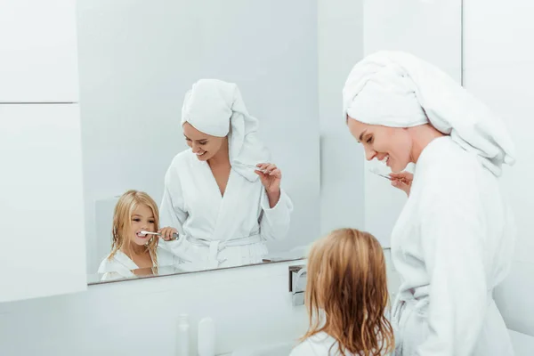 Happy mother looking at daughter brushing teeth near mirror — Stock Photo