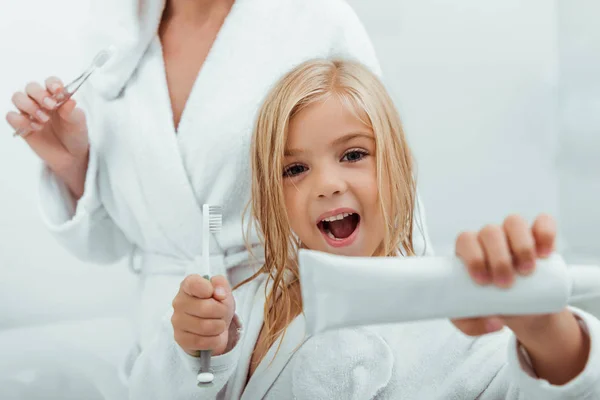 Enfoque selectivo de niño alegre sosteniendo pasta de dientes y cepillo de dientes cerca de la madre - foto de stock