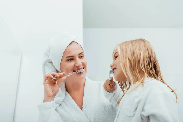 Happy kid brushing teeth near cheerful mother in bathrobe — Stock Photo