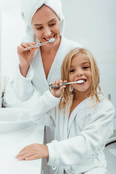 Feliz niño cepillarse los dientes cerca atractiva madre en albornoz - foto de stock