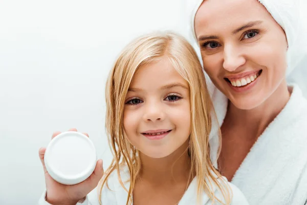 Criança bonito segurando recipiente com creme cosmético perto mãe alegre em roupão de banho — Fotografia de Stock