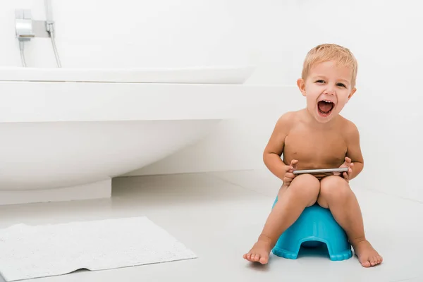 Excited toddler boy with opened mouth sitting on blue potty and holding smartphone near bathtub — Stock Photo