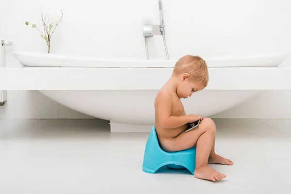 Vue latérale du garçon tout-petit assis sur le pot bleu et tenant smartphone près de la baignoire — Photo de stock