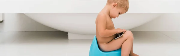Plan panoramique de tout-petit garçon assis sur le pot bleu et tenant smartphone près de la baignoire — Photo de stock